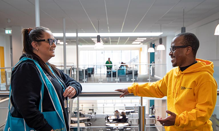 Two students talking whilst stood on a balcony inside a building