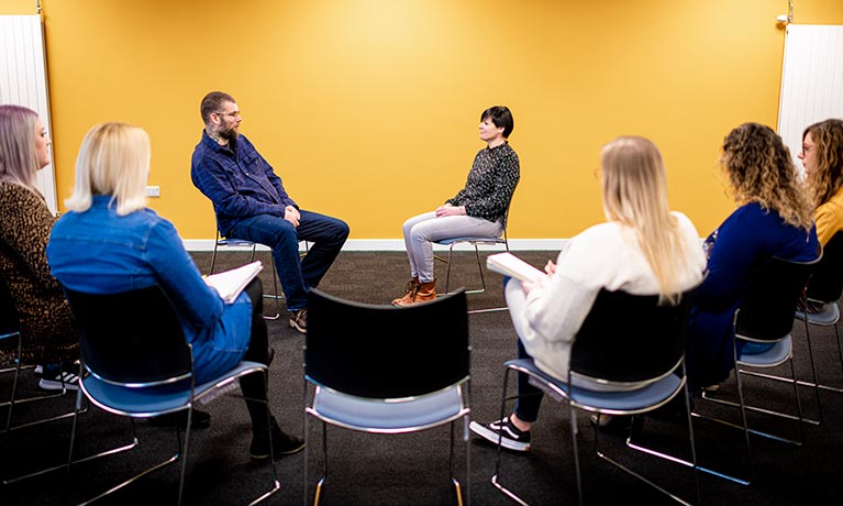 Two students sat at the front of a room with other people watching