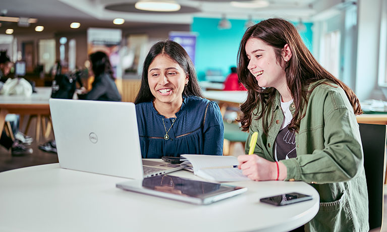 Two students sat at a looking at a laptop and writing 