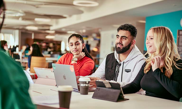 Group of students sat at a table with their laptops and tablets talking