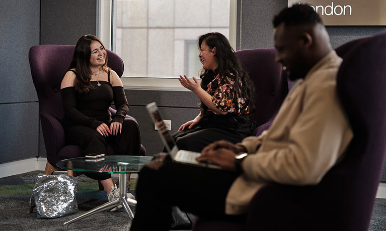 Two ladies smiling and sitting on purple armchairs and one man typing on a laptop