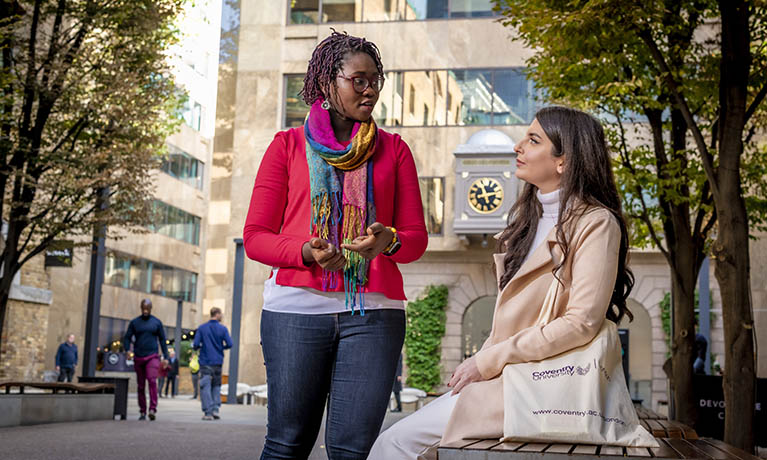 two young females chatting outside on a tree lined street 