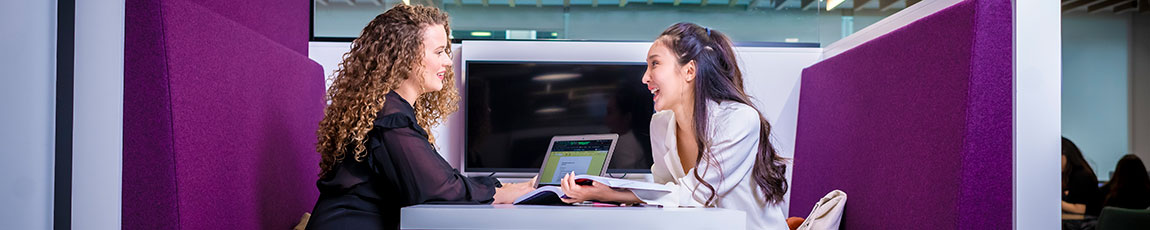 Two ladies sitting on purple sofas chatting in front of an open laptop