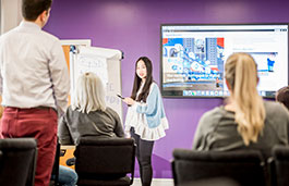 lady standing up presenting to three people  using a flip chart and presentation on screen