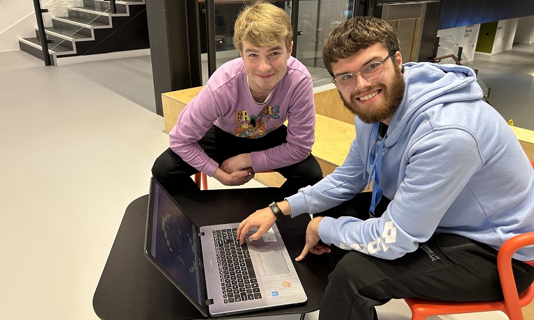 Students William and Sam sat in chairs next to a table on which sits a laptop with 8-Bit Rainforest on it