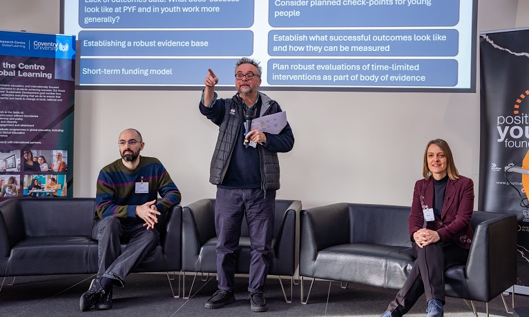 Three people on a stage looking past the camera with a presentation behind them on a screen