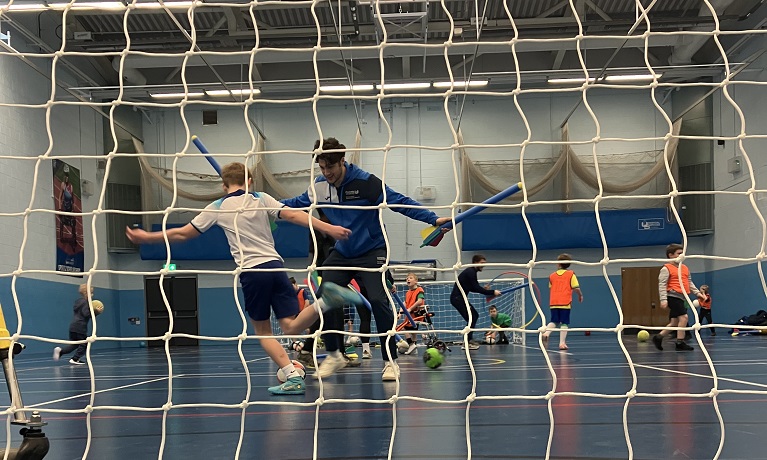 Children playing football in a sports hall with Coventry University staff chasing them while holding foam javelins