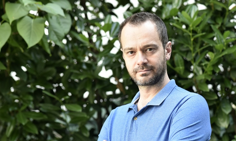 Head and shoulders image of Federico Carollo in a blue t-shirt with green leaves in the background