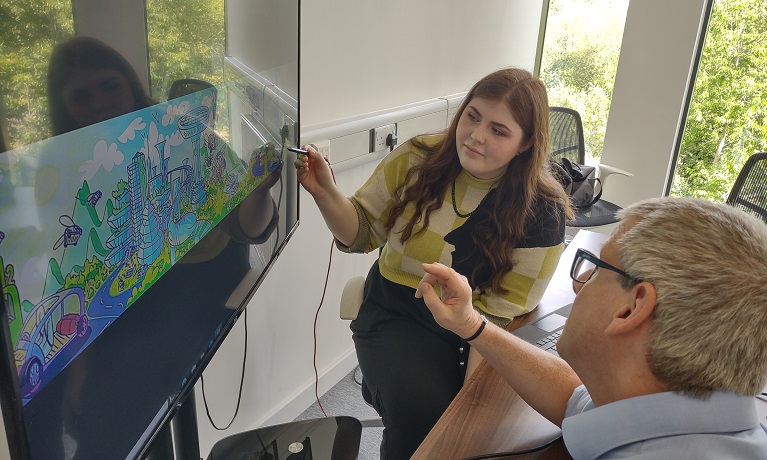 A picture of a female student sitting next to a display screen with a man