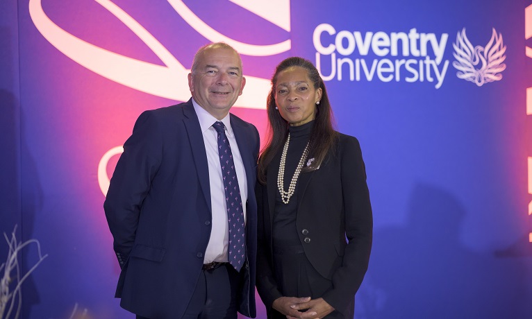 Coventry University Vice-Chancellor John Latham and Chancellor Margaret Casely-Hayford stood infront of a university logo and smiling at the camera