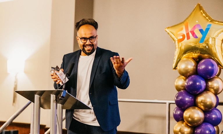 Danny Mirza on stage collecting his award with purple and gold balloons in the background