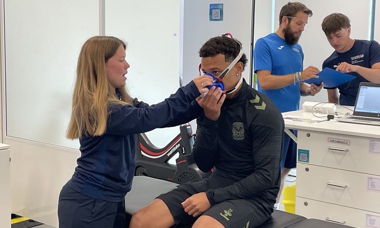 Coventry City player Joel Latibeaudiere being helped to wear a mask by a Coventry University student