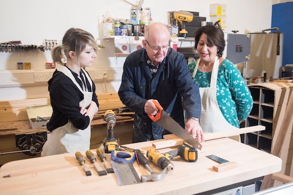 Middle-aged and young adult watch a senior citizen cut wood with a hand saw