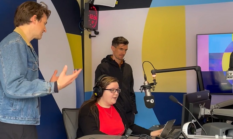 A female student sat at a radio desk with two male Radio 1 staff members standing either side of her