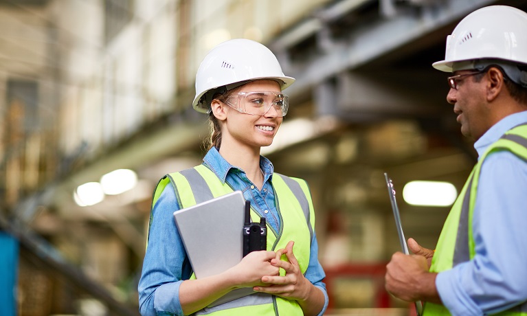 A young woman wearing a hi-vis jacket and a hard hat holding walkie talkie smiling at a man also wearing a hi-vis jacket and hard hat who is also holding a clipboard