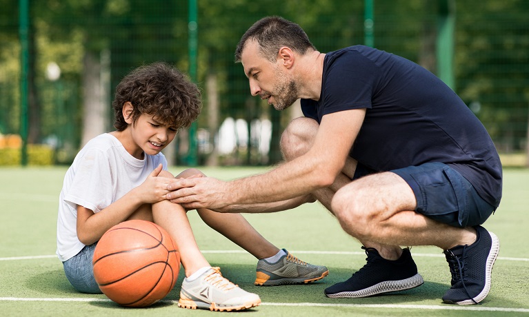 A boy sitting on the floor holding his knee while a man leans down to help him and a basketball sits motionless next to the boy