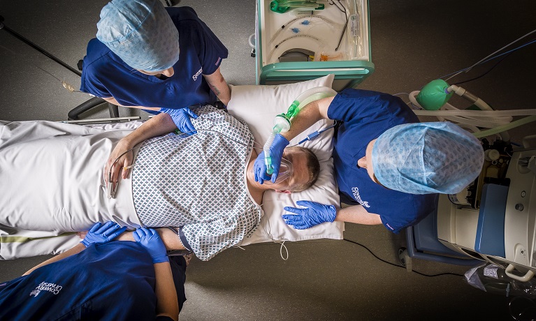 An image from above of three doctors in Coventry University uniforms around a male patient lying on a bed with a breathing mask being held over his face