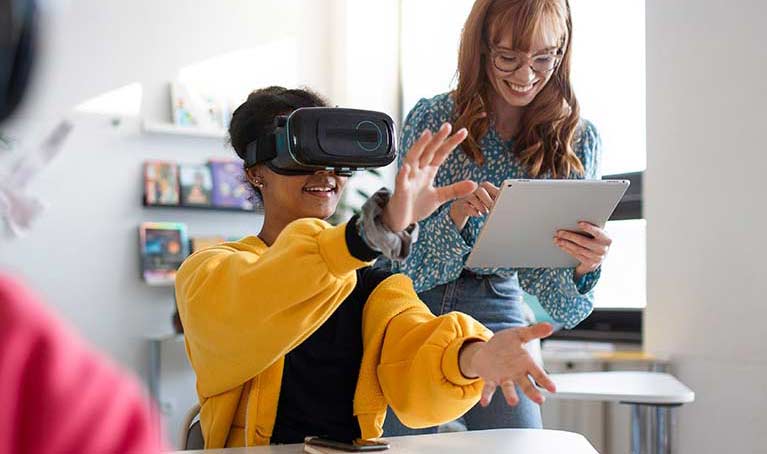 Teacher in a class room holding a tablet standing beside a girl wearing a VR headset