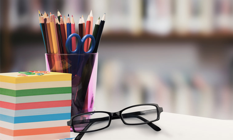Glasses, post-it notes and coloured pencils in a pot on a desk with a bookcase in the background