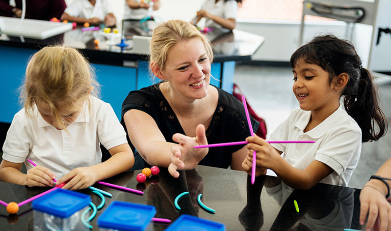 A teacher with blonde hair kneeling down next to a student using chemistry molecular models.