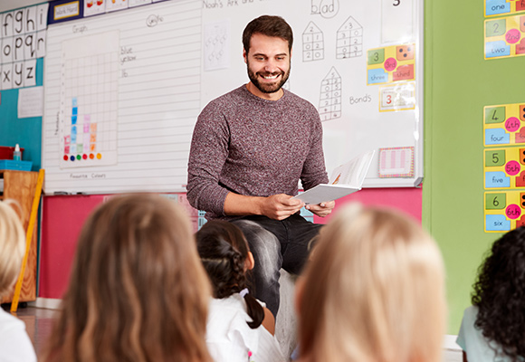A teacher stood up in front of the classroom, with drawings and the alphabet on the wall behind him.