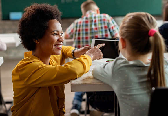 A teacher kneeling down to a young student's desk, showing her how to use a tablet.