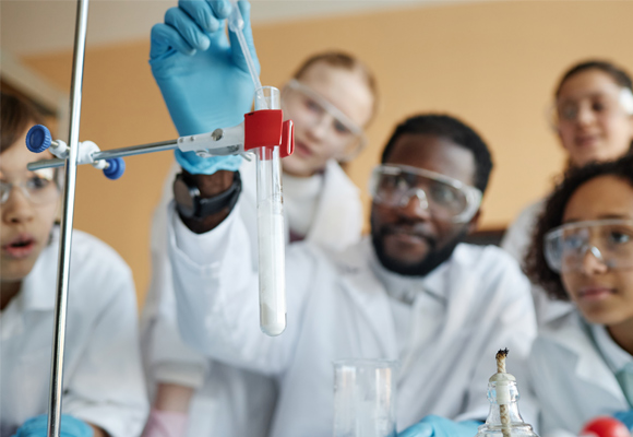 A teacher in a lab coat and safety glasses dropping liquid into a test tube, watched by schoolchildren in lab coats and safety goggles.