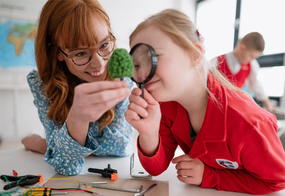 A teacher with holding up a tree model while a young student with Down syndrome holds up a magnifying glass to it.