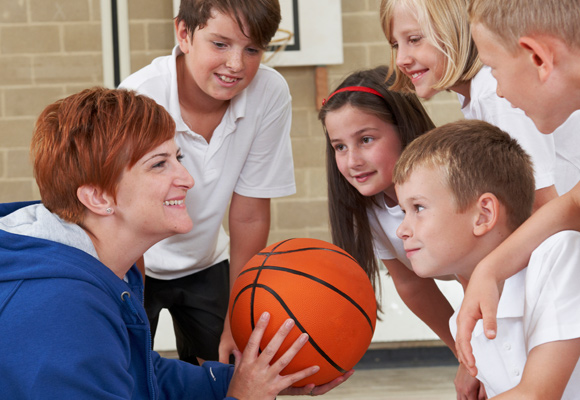 A teacher holding a basketball with a team of primary students in PE class.