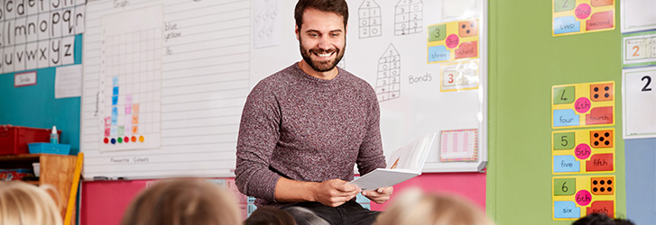 A male teacher smiling at the front of a classroom of young schoolchildren.
