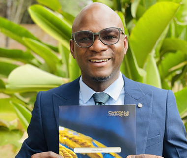 Chilipo Gunda outdoors against a background of tropical plants, holding a graduation certificate from Coventry University