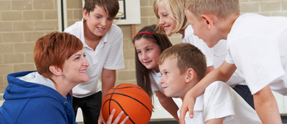 A teacher holding a basketball in a school sports hall, giving a team talk to a group of primary school-aged children