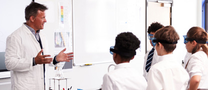 A teacher in a lab coat with a flask on the desk in front of him, talking to a group of students wearing safety glasses
