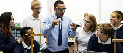 A teacher in a chemistry lab holding a test tube up in front of students wearing safety glasses