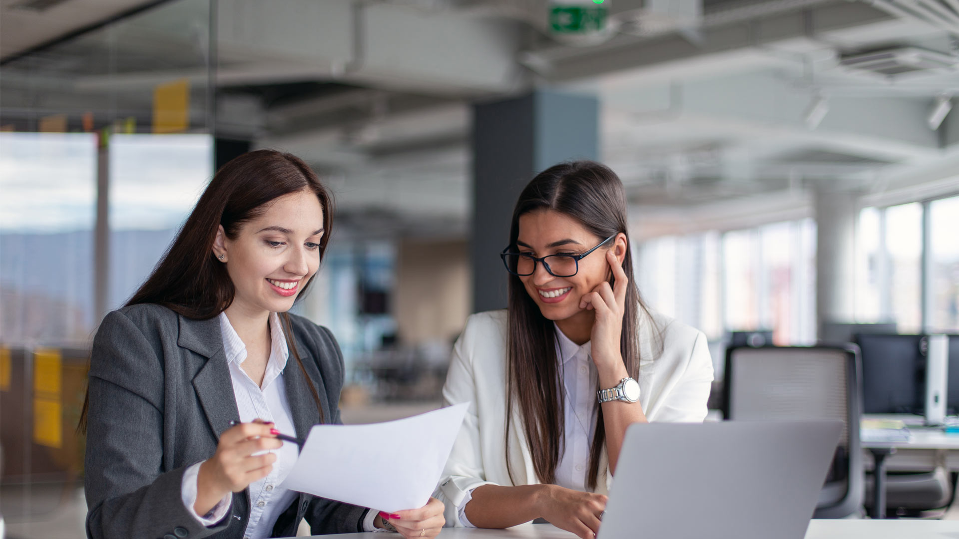 Two women in professional workwear at a desk with a laptop, looking over some papers and smiling.