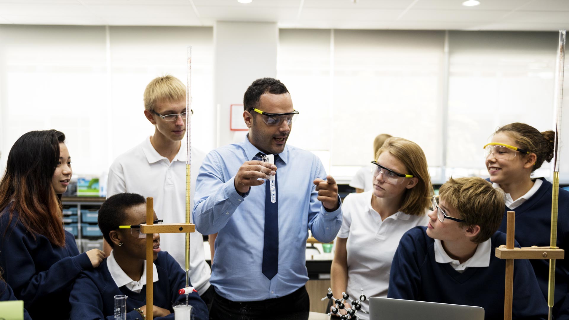 A teacher in a chemistry lab holding a test tube up in front of students wearing safety glasses.