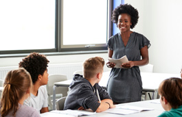 A teacher stood in front of a group of students sat around a large table.