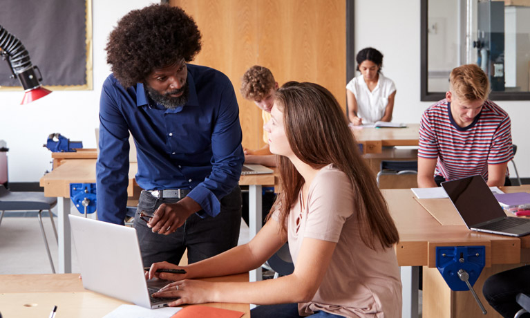 A teacher talking to a student who is working on a laptop.