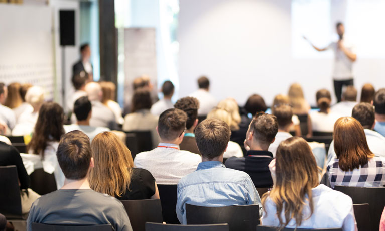 A shallow depth of field focus on an audience and lecturer standing at the front of the room