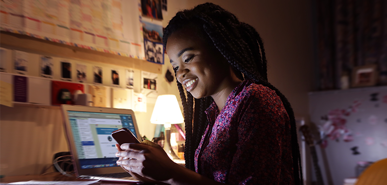 Black female student working on a laptop and looking at her phone in her bedroom