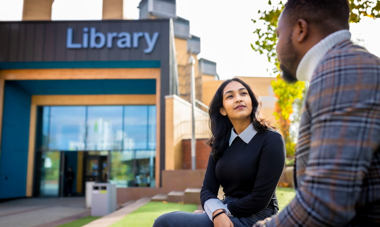 Two students sat on a wall outside in front of the library